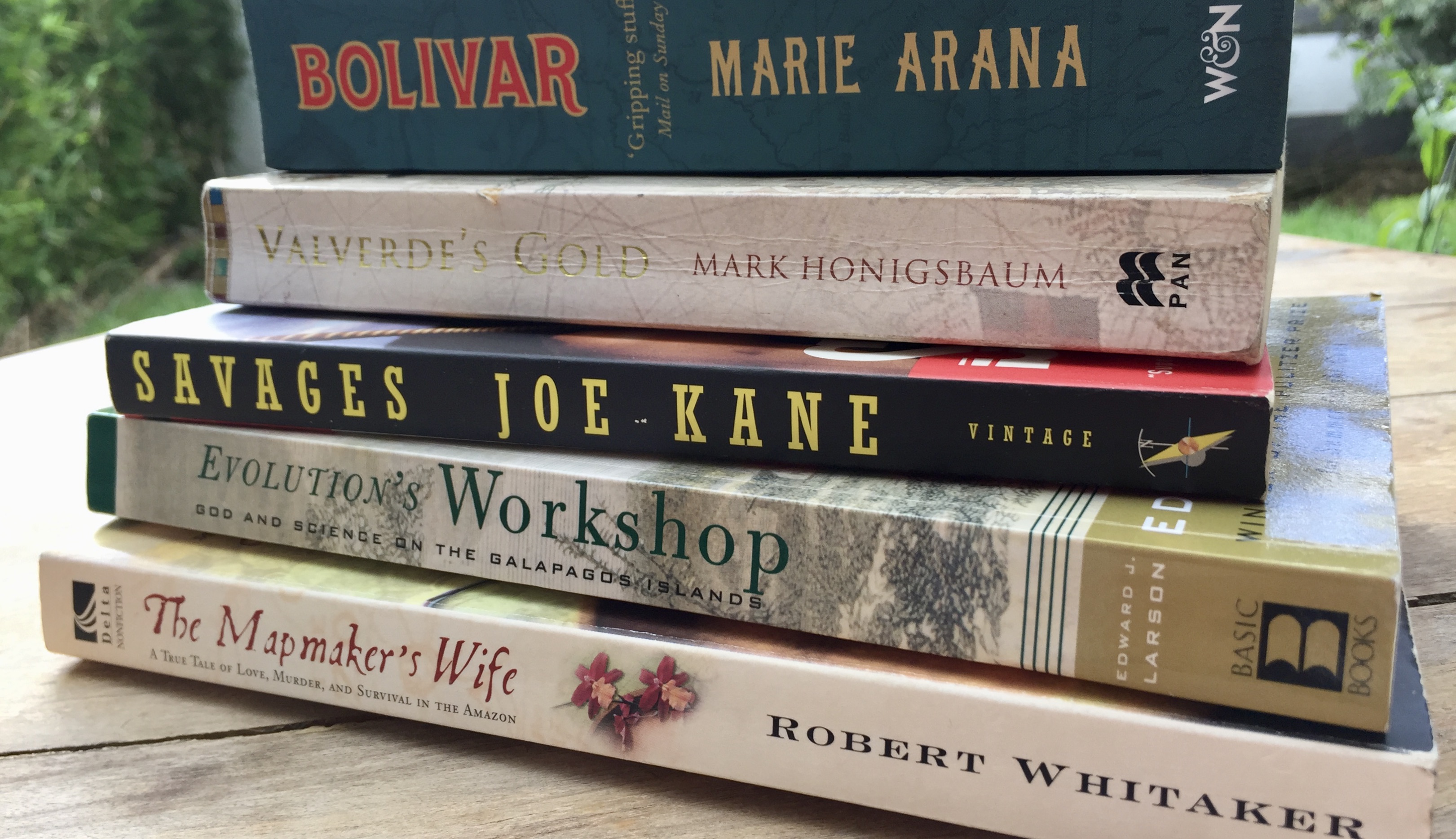 Five books lying on their sides, one on top of the other, on a wooden table in a garden.