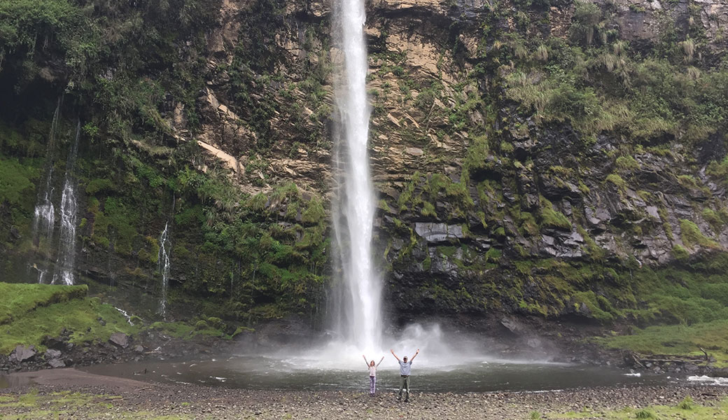 Dad and daughter with arms in air in front of tall Condor Machay Waterfall in Cotopaxi